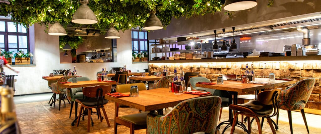 Intérieur restaurant, tables bois, chaises colorées, feuillage suspendu lumineux, cuisine ouverte avec mur pierre, personne marche.