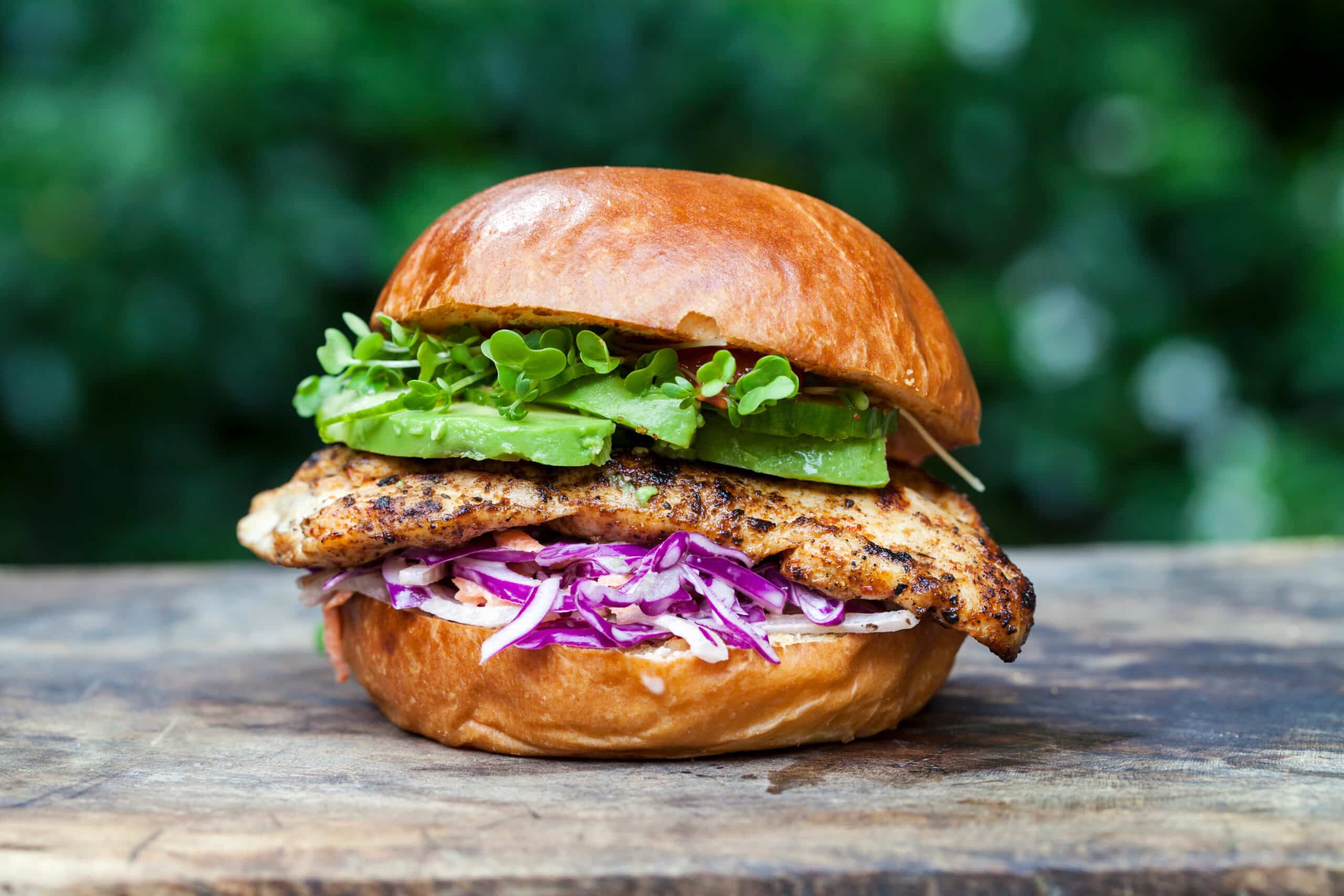 Burger de poulet grillé avec avocat, coleslaw rouge et pousses vertes sur un pain brioché.